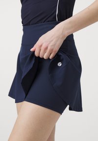 Navy blue athletic skort with a flared design, made from textured fabric, featuring a smooth waistband and a logo on the side.