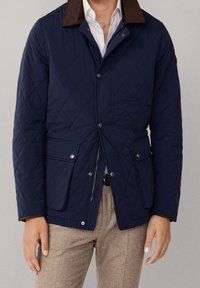 Homme portant une veste matelassée bleu marine avec col marron, chemise blanche et pantalon beige, debout devant un fond neutre.