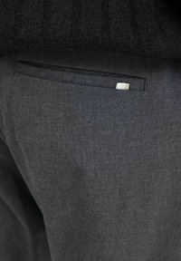 Dunkelgraue Hosen mit einer dezenten Textur, einer standardmäßigen Gesäßtasche mit einem gestreiften Stoffakzent. Der Bund hat keine sichtbaren Verschlüsse.