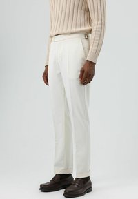 Witte, op maat gemaakte broek met een gladde textuur, voorzien van een zijdelingse verstelknop, gecombineerd met donkerbruine loafers en een lichtbeige geribbelde trui.