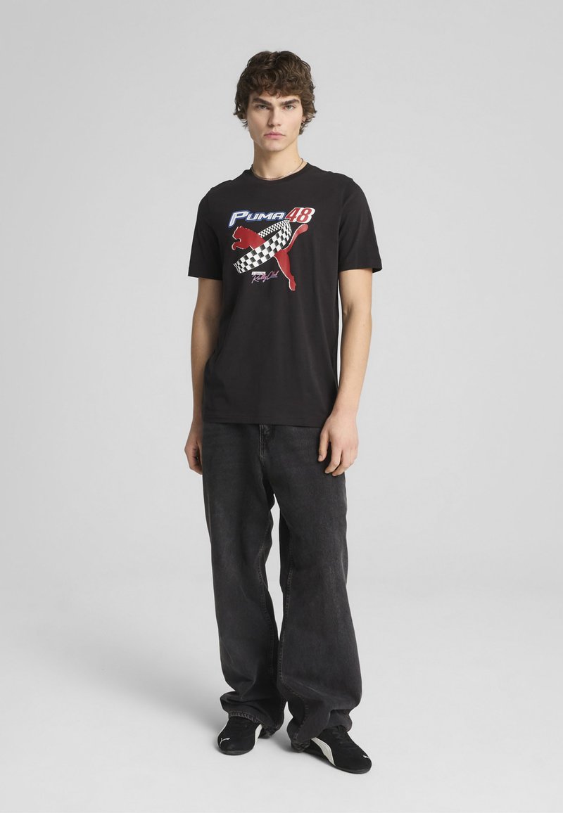 T-shirt en coton noir avec un design graphique comportant un puma rouge et un motif de drapeau à damier, associé à un jean foncé à coupe ample et des chaussures noires.