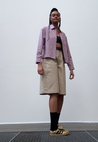 Helles lila gemustertes Hemd, schwarzes Crop-Top, beige Shorts, schwarze Socken und goldene Sneakers mit schwarzen Streifen. Einfaches, lässiges Outfit.