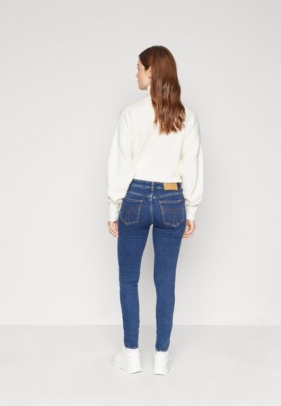 Jean ajustados azules de cintura alta, con bolsillos traseros y costuras claras, combinados con un suéter crema y zapatillas blancas.