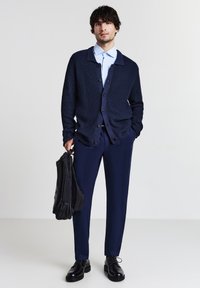 Uomo in cardigan blu navy, camicia azzurra, pantaloni navy, scarpe nere che tiene uno zaino nero contro uno sfondo chiaro uniforme.