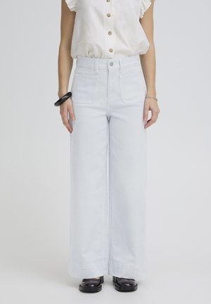 Sieviete, kas valkā gaismazilas platām svītrām dekorētas bikses ar augstu vidukli, baltu bezrokas pogājamu topu, melnus masīvus apavus un rokassprādzes uz abām plaukstām.