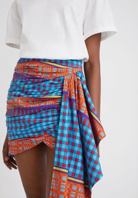 Värikäs kuviollinen hame, joka on valmistettu kankaasta, jossa on sinisiä, oransseja ja punaisia ruudullisia kuvioita. Hameessa on rypytetty tekstuuri ja solmittava koriste.