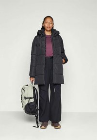 Giubbotto imbottito nero con cappuccio, caratterizzato da un design trapuntato. Abbinato a pantaloni neri a gamba larga, una maglietta viola e uno zaino verde chiaro.