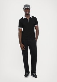 Svart pikétrøye med glidelåskrage og hvite mønstrete mansjetter. Kombinert med svarte bukser og svarte sneakers, komplettert med en grå caps.