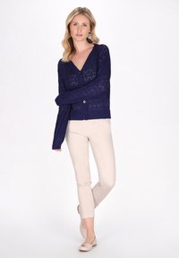 Marinblå stickad kofta med V-ringning, mönster med öppningar och långa ärmar; ihopparad med beige figursydda byxor och beige ballerinas.