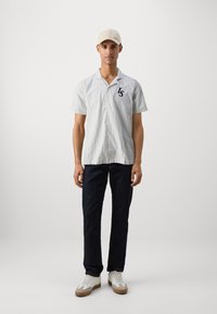 Camisa de manga corta a rayas en azul claro y blanco, con cuello y logo bordado en negro. Combinada con jeans oscuros y zapatillas blancas.