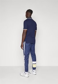 Marineblå polo skjorte med hvit og gull aksentbånd, kombinert med marineblå joggebukser med hvite og gule sidestiper. Hvite sneakers.