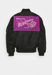 Black bomber jacket featuring a large, textured purple patch with white text: "MAGIC FOR THE RESISTANCE." Ribbed cuffs and collar.