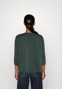 Olivgrüne Bluse mit glatter, glänzender Textur, Drei-Viertel-Ärmeln und geradem Schnitt, kombiniert mit dunkelblauen Jeans.