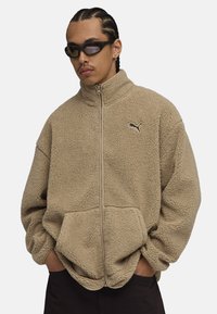 Chaqueta de forro polar beige con cuello alto, cierre de cremallera y bolsillos laterales; presenta un pequeño logo negro en el pecho. Material suave y texturizado.