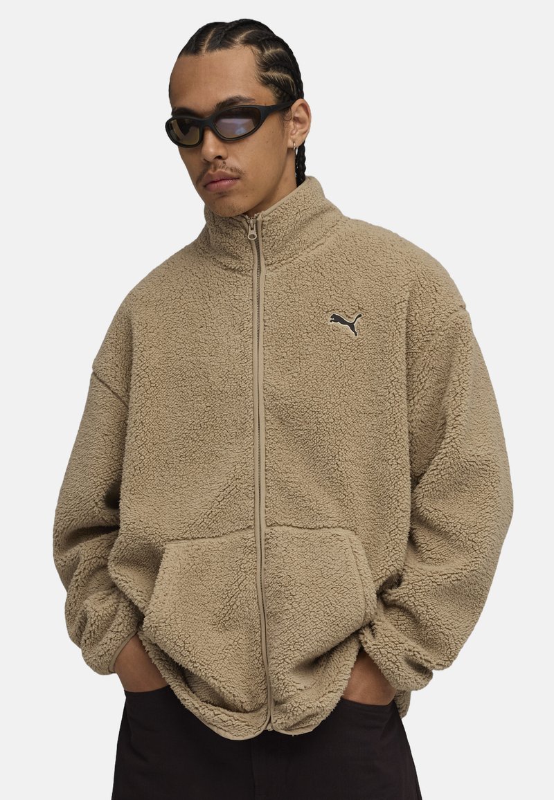Chaqueta de forro polar beige con cuello alto, cierre de cremallera y bolsillos laterales; presenta un pequeño logo negro en el pecho. Material suave y texturizado.