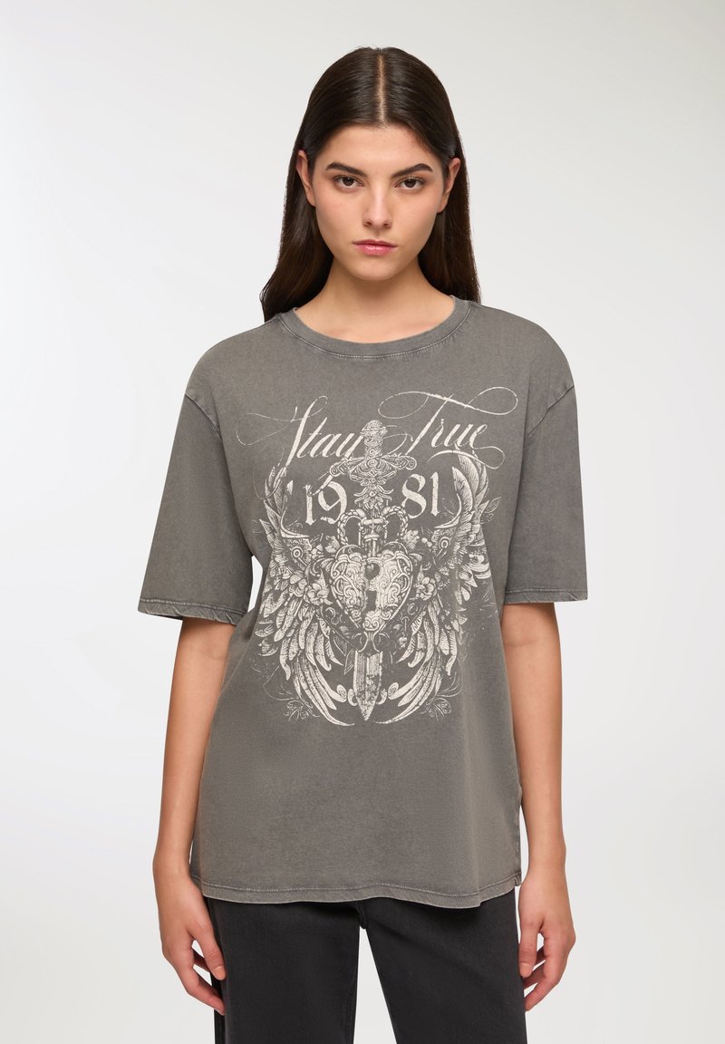 T-shirt grigio oversize con una texture sbiadita, caratterizzata da una grafica bianca di ali e un cuore con il testo "Stay True 1981" in un carattere decorativo.