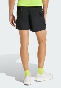 Man wearing black athletic shorts, bright yellow socks, white running shoes, standing with back facing camera on a light background.