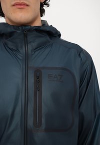Marine vandtæt jakke med en lynlåslomme på brystet. Har hætte og glat overflade, med brandlogo og minimalistiske designelementer.
