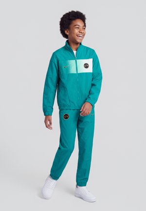 Jeune personne souriante debout, portant un survêtement Nike bleu sarcelle avec des chaussures blanches sur un fond gris clair uni.