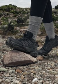 Scarpe da ginnastica nere con tomaia testurizzata e suole robuste, dotate di un logo sul tallone, posizionate su una grande roccia in un terreno roccioso.