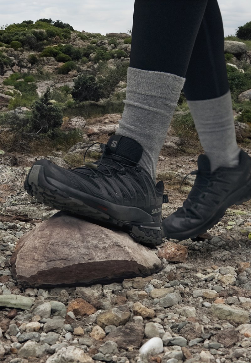 Scarpe da ginnastica nere con tomaia testurizzata e suole robuste, dotate di un logo sul tallone, posizionate su una grande roccia in un terreno roccioso.