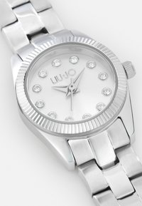 Montre en métal argenté avec une lunette striée et un cadran blanc. Elle dispose de douze marqueurs en cristal, d'un bracelet en métal et d'un mouvement simple à trois aiguilles.