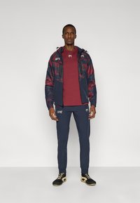 Αθλητική στολή σε ναυτικό μπλε και μαρόν, που περιλαμβάνει μια ζακέτα με καμουφλάζ μοτίβο, παντελόνια που ταιριάζουν και μαύρα παπούτσια με χρυσές λεπτομέρειες.