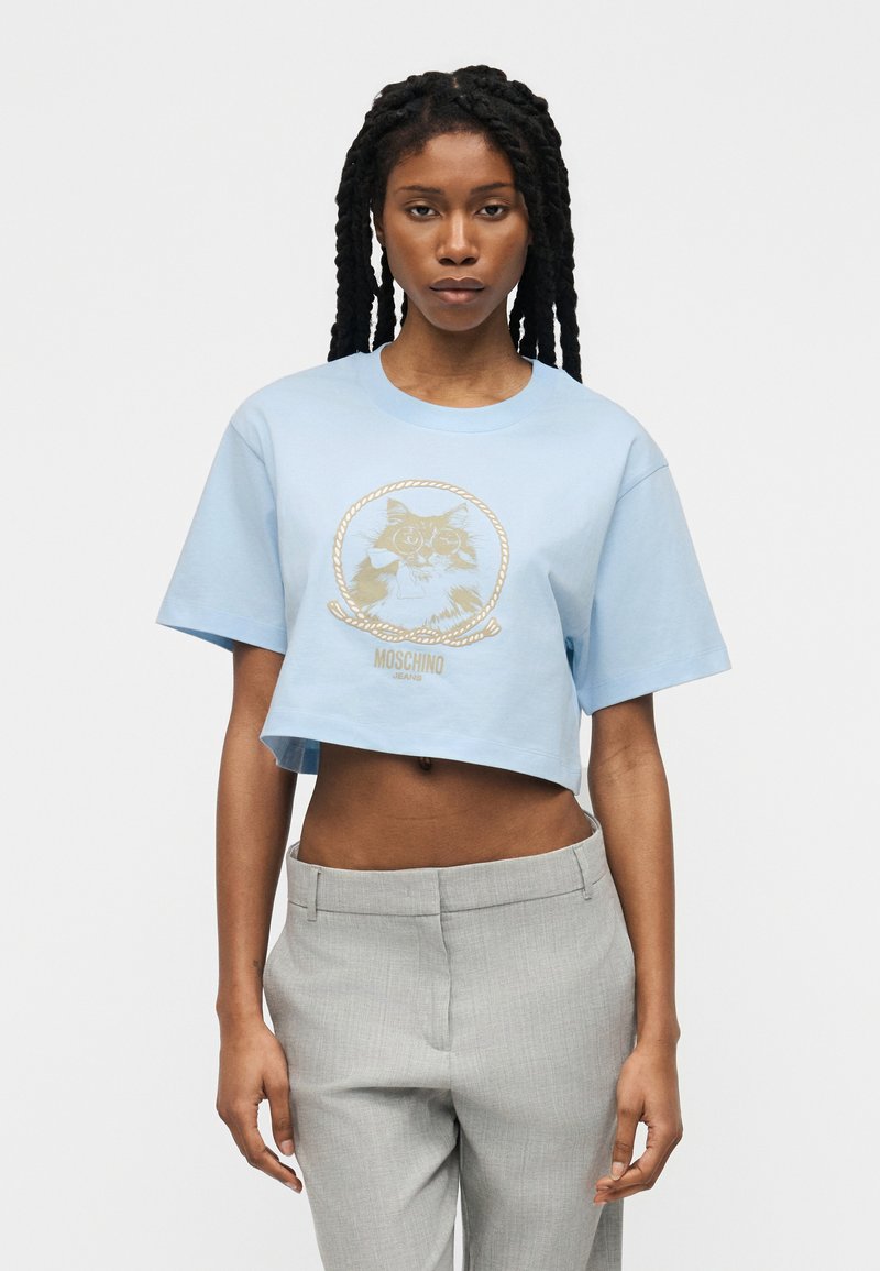 Γυναίκα με μακριά πλεξούδα που φοράει ένα ανοιχτό γαλάζιο κοντό μπλουζάκι με γραφικό γάτας και γκρι ψηλόμεσα παντελόνια, στέκεται μπροστά σε ένα απλό φόντο.