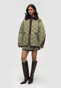 Kvinde iført en oversized, quilted olivengrøn jakke og matchende nederdel, parret med høje, mørkebrune læderstøvler, stående mod en ensfarvet baggrund.