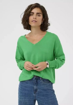 Femme aux cheveux bruns courts portant un pull vert à col en V, un jean bleu, un bracelet argenté et des boucles d'oreilles créoles sur un fond uni.