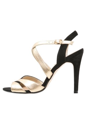 Musta mokkanahka ja kultainen metallinen remmisaapas korkean stiletto-kannan kanssa, jossa on ristikkäiset remmit ja solki nilkassa.