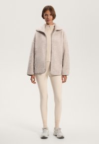 Blød beige jakke med en fluffy tekstur, høj krave og lynlås, båret over en lys beige top og tætsiddende leggings, parret med sneakers.