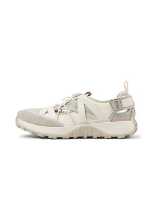 Beige und cremeweiße sportliche Sandale mit dicker Gummisohle, gekreuzten Stoffriemen, verstellbarem Riemen hinten und Schnürverschluss vor weißem Hintergrund.