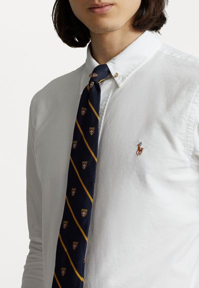Witte katoenen overhemd met een button-down kraag, gecombineerd met een marineblauwe stropdas met gele diagonale strepen en kleine schildpatronen.