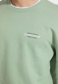 Bomba mint zelena iz bombaža, z okroglim izrezom, teksturirano tkanino in belim vezenim logotipom: "GUESS JEANS" in "AMERICAN TRADITION."