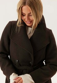 Blondynka w ciemno-brązowym płaszczu z paskiem i białym swetrze, patrzy w dół i delikatnie się uśmiecha.