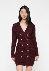 Abito a maglia bordeaux con tessuto testurizzato, scollatura a V, maniche lunghe e bottoni dorati doppi. Silhouette aderente con dettagli a coste.