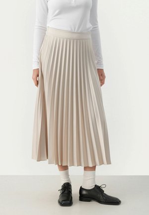 Beige Midirock mit Falten, breitem Bund und fließendem Design. Getragen mit schwarzen Schuhen und weißen Socken. Glatte Textur und weicher Fall.