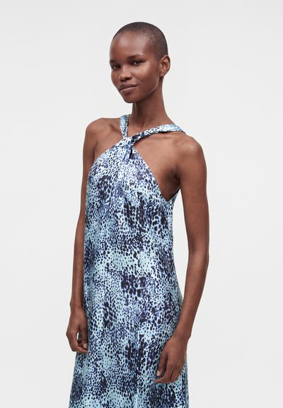 Femme aux cheveux courts portant une robe sans manches bleue et noire à pois avec un décolleté halter torsadé sur un fond clair uni.