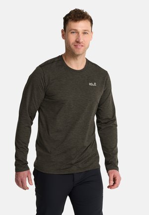 Uomo con una maglietta Jack Wolfskin a maniche lunghe verde scuro e pantaloni neri in piedi davanti a uno sfondo chiaro e uniforme.