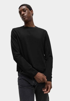 Man die de manchette van een zwart shirt met lange mouwen aanpast, draagt donkergrijze jeans, staand tegen een effen achtergrond.