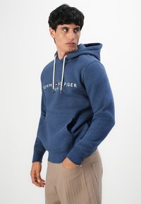 Man in een blauwe Tommy Hilfiger hoodie en beige broek, staand met één hand in de zak, kijkend opzij.