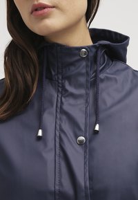 Kvinna i mörkblå regnkappa med hög krage, huva och dragsnöre med metalldetaljer, läpparna något frånskilda.