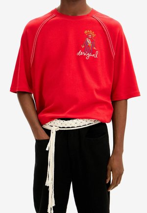 Personne portant une chemise rouge à manches courtes avec une figure brodée colorée et du texte, un pantalon noir et une ceinture blanche en corde tressée.