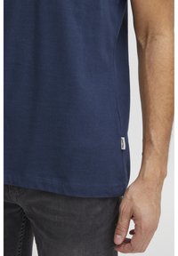 T-shirt di cotone blu navy con maniche corte, caratterizzata da un orlo dritto e una piccola etichetta del marchio sul lato, abbinata a pantaloni grigio scuro.