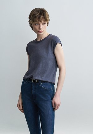 Jeune personne aux cheveux courts et bouclés portant un t-shirt gris foncé aux manches retroussées, rentré dans un jean bleu taille haute avec une ceinture noire.