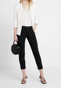 Kobieta w białym żakiecie i koszuli, czarnych spodniach 3/4, czarnych sandałach na obcasie, trzymająca okrągłą czarną torebkę, pozująca na białym tle.