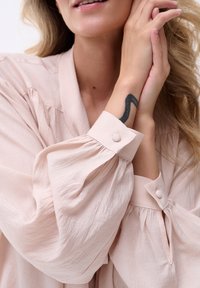 Jasnoróżowa bluzka z bufiastymi rękawami, z guzikami przy mankietach i miękkim, teksturowanym materiałem. Projekt zawiera fałdy oraz luźny krój.