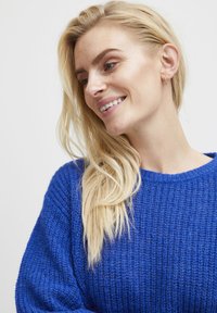 Pullover blu brillante lavorato a maglia con una vestibilità ampia, tessuto texturizzato e scollatura larga. Lunghi capelli biondi e mossi si adagiano parzialmente su una spalla.