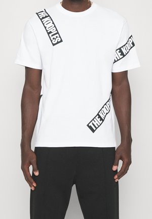 Man draagt een wit T-shirt met korte mouwen met zwarte diagonale tekst "THE KOOPLES" en effen zwarte broek tegen een neutrale achtergrond.